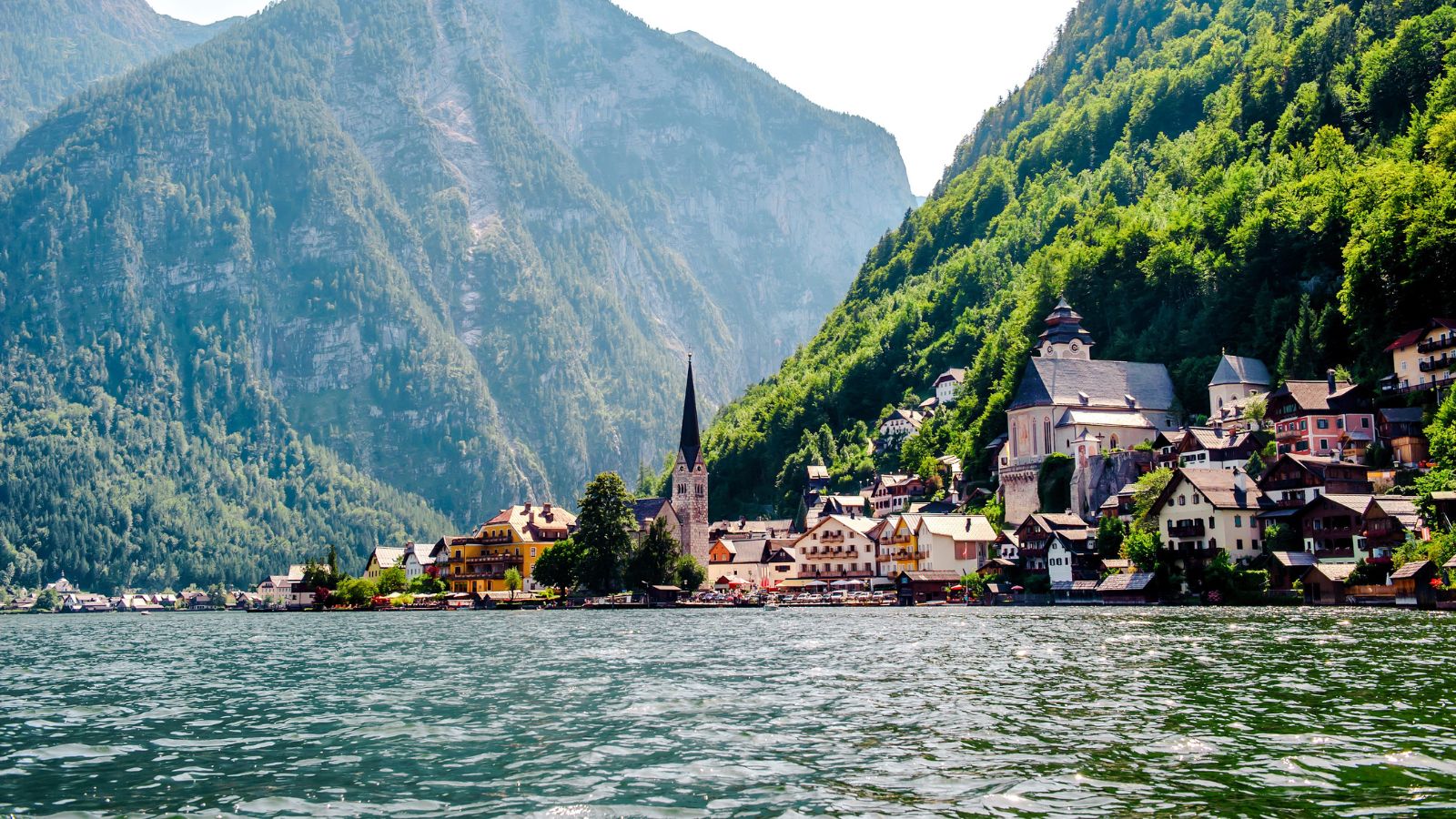 A lakeside village with a church steeple and traditional houses sits below forested mountains under a bright sky.