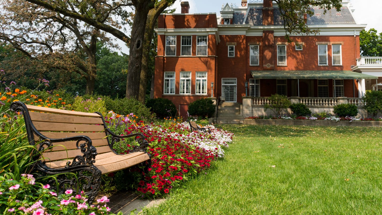 A wooden bench is next to a vibrant flowerbed before a large brick house with a covered porch and tidy lawn.