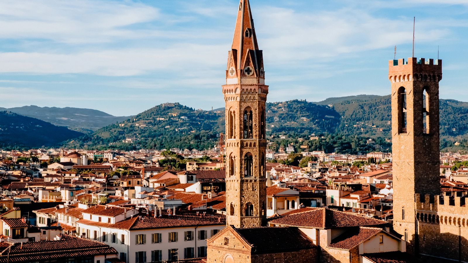 Two stone towers rise above red-roofed buildings in a historic city, with green hills and blue sky in the background.