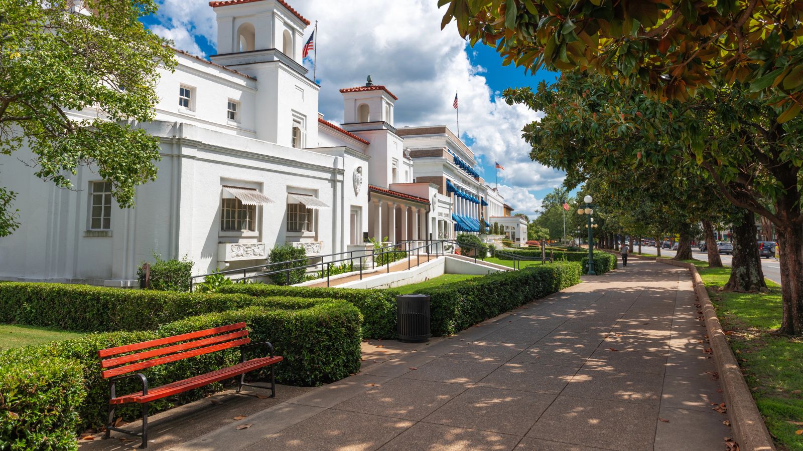 A sidewalk borders a trimmed hedge and red bench near a white building and trees beneath a partly cloudy sky.