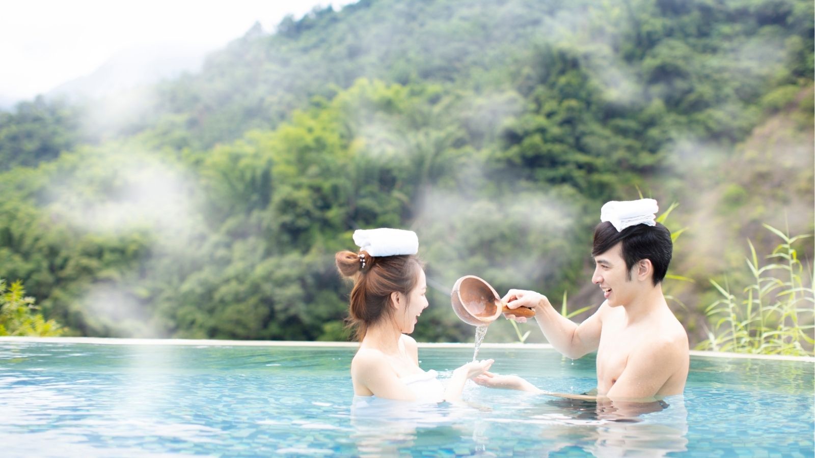 Two people in a pool with towels on their heads, one pouring water from a ladle onto the other's hand, with green hills behind.