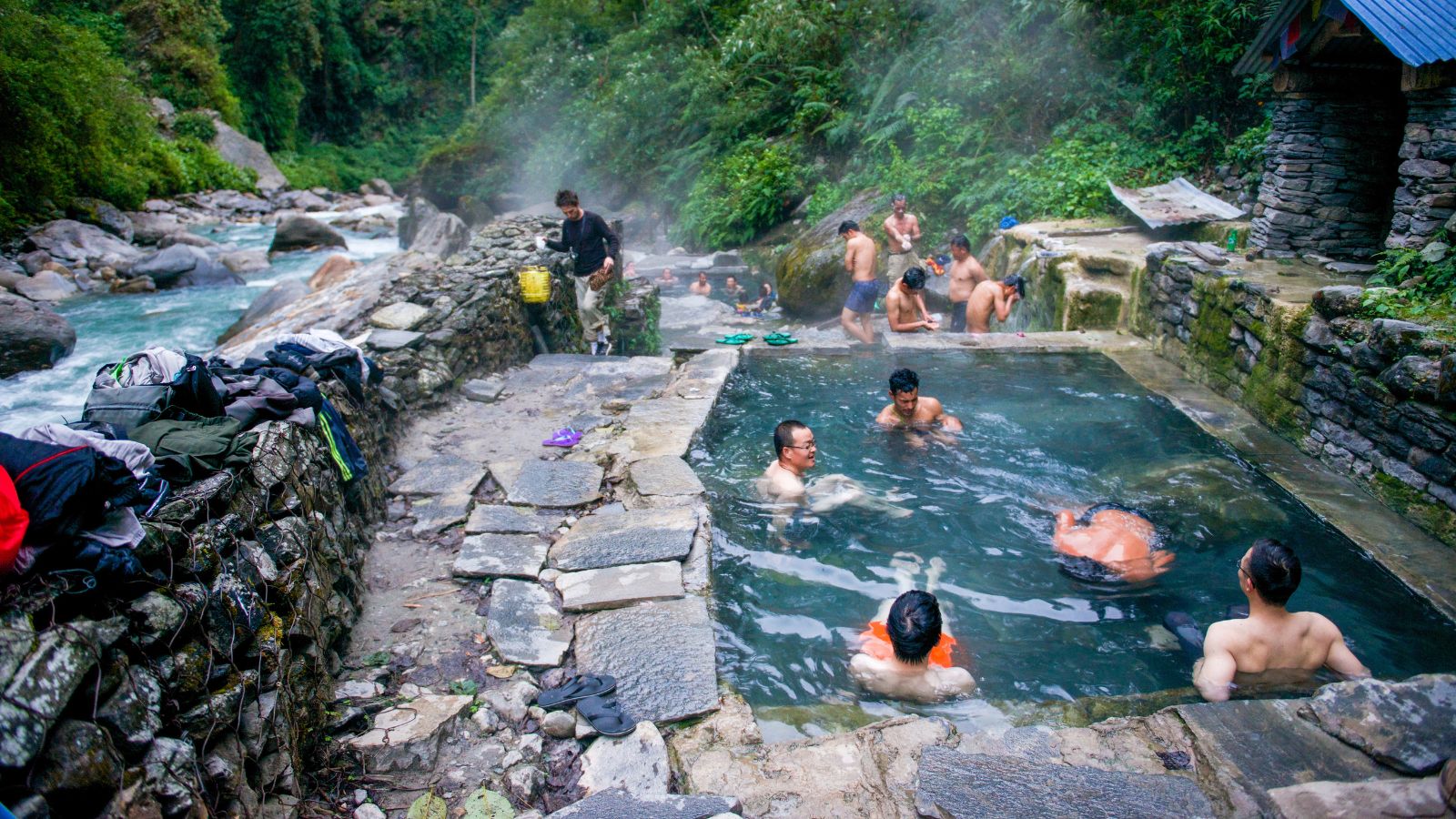 People bathe in an outdoor hot spring pool bordered by stone walls and greenery, with clothes laid on a nearby stone ledge.