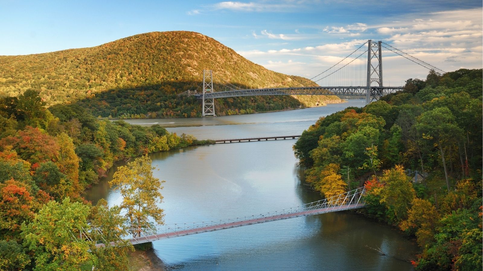 A suspension bridge spans a wide river backed by autumn forests, with a smaller footbridge visible in the foreground.