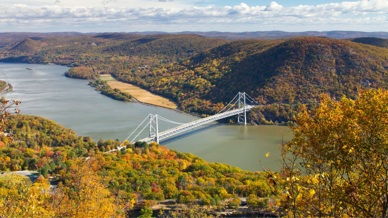 A suspension bridge crosses a wide river, bordered by autumn-colored forested hills beneath a partly cloudy sky.