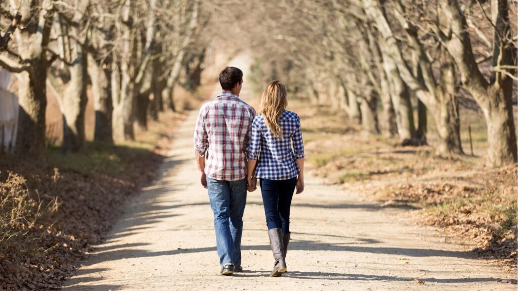A man and woman in plaid shirts and jeans walk hand in hand down a dirt path lined with leafless trees.