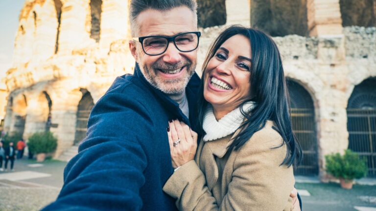 A smiling man and woman take a selfie in front of an ancient stone building with arched openings.
