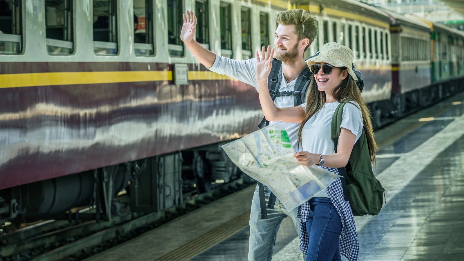 A man and woman wave and hold a map on a train platform, with a train in the background.