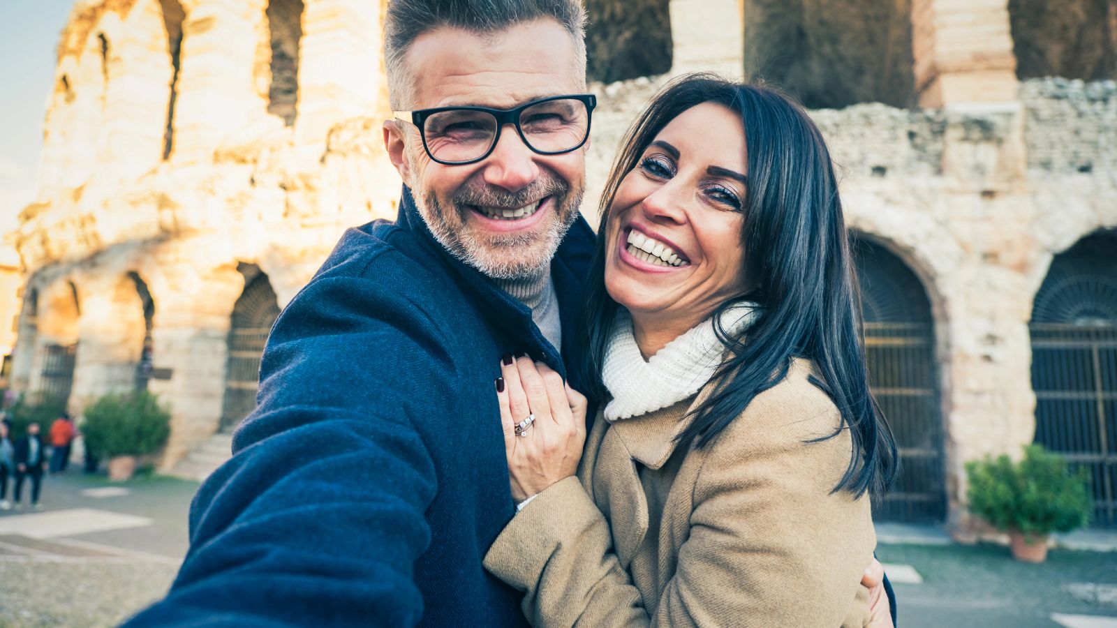 A smiling man and woman take a selfie in front of an ancient stone building with arched openings.