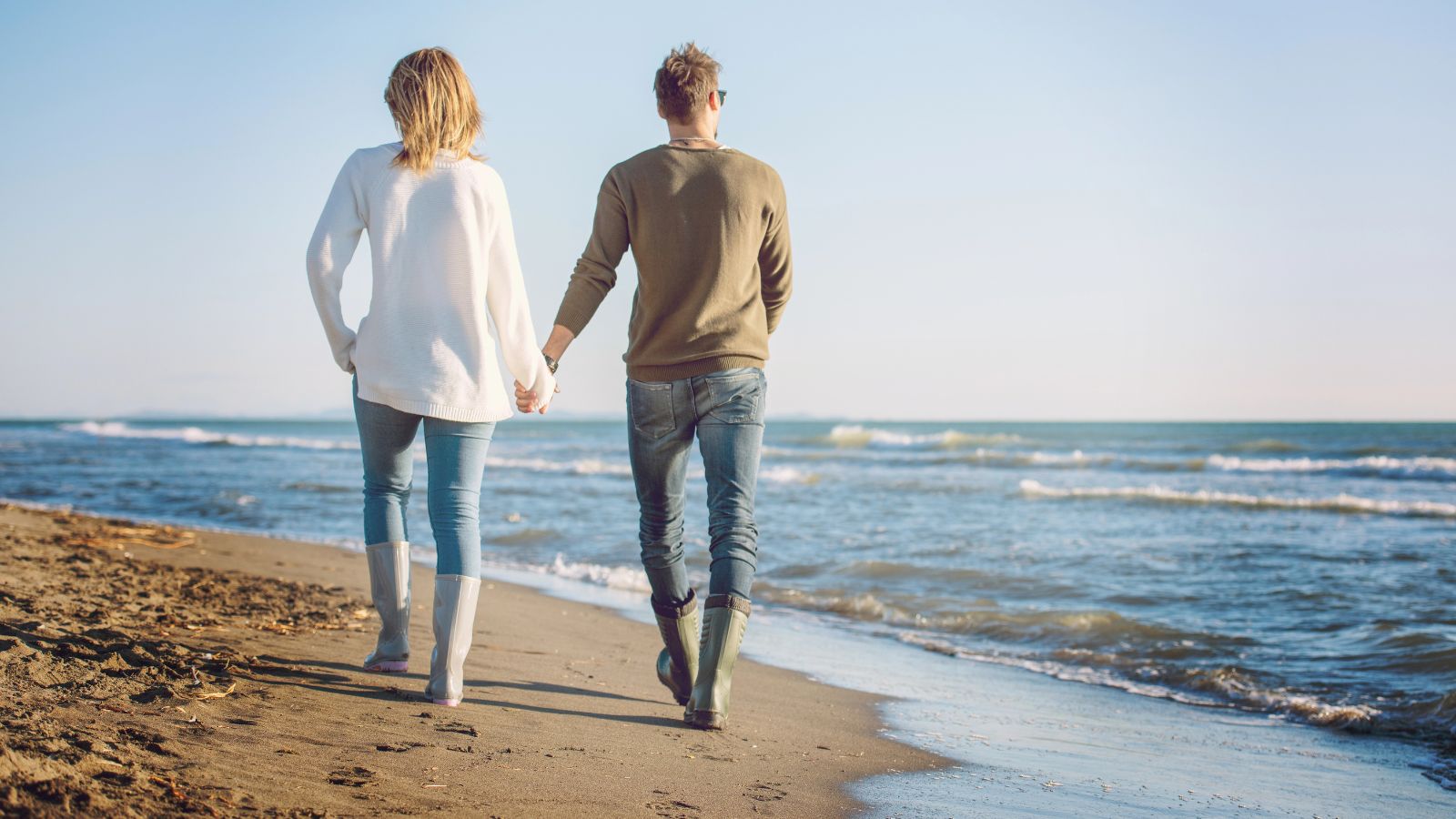 Two people hold hands while walking on a sandy beach by gentle waves under a clear sky.