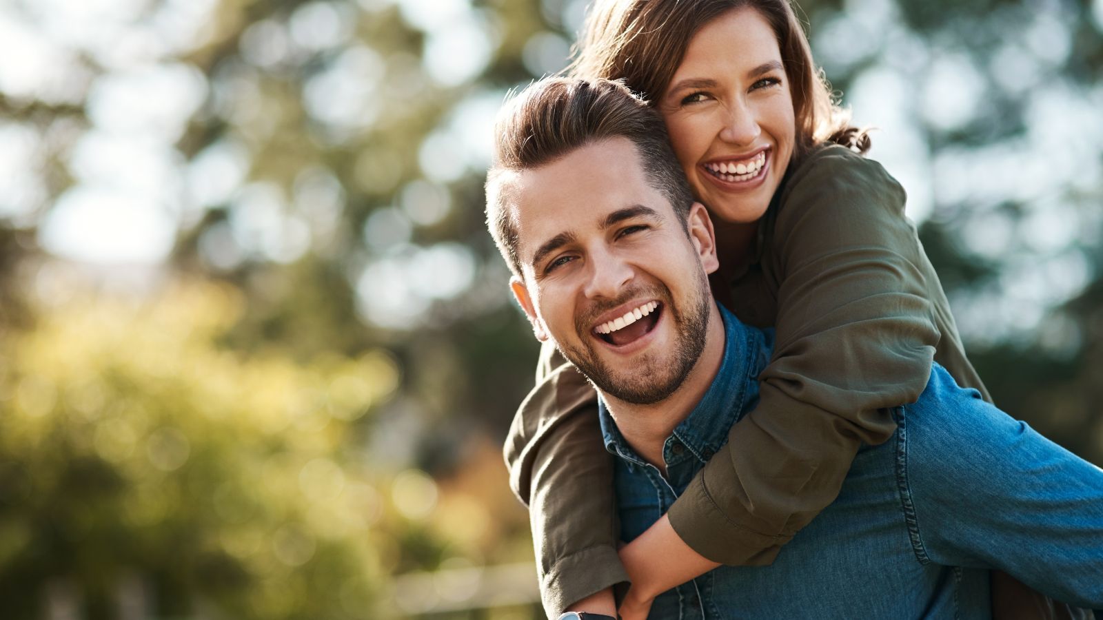 A woman gives a piggyback ride to a laughing man outdoors, both smiling at the camera against a backdrop of blurred greenery.