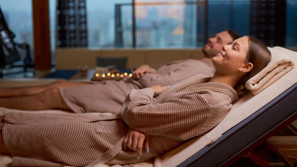 Two people in brown robes lounge at a spa, candles glowing in the background.
