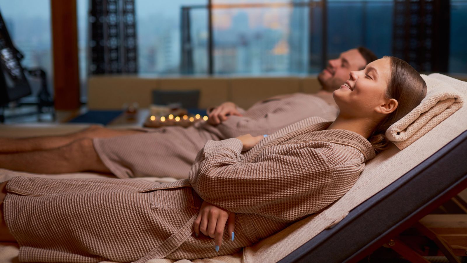 Two people in brown robes lounge at a spa, candles glowing in the background.