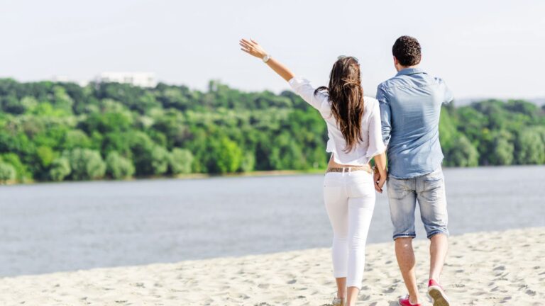A man and woman walk hand-in-hand along a sandy beach by the water, with green trees in the background.