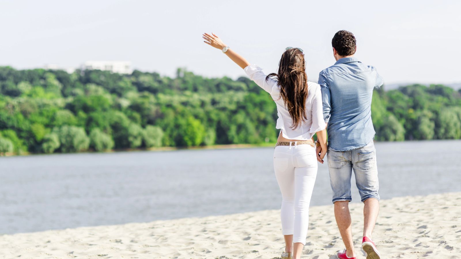 A man and woman walk hand-in-hand along a sandy beach by the water, with green trees in the background.