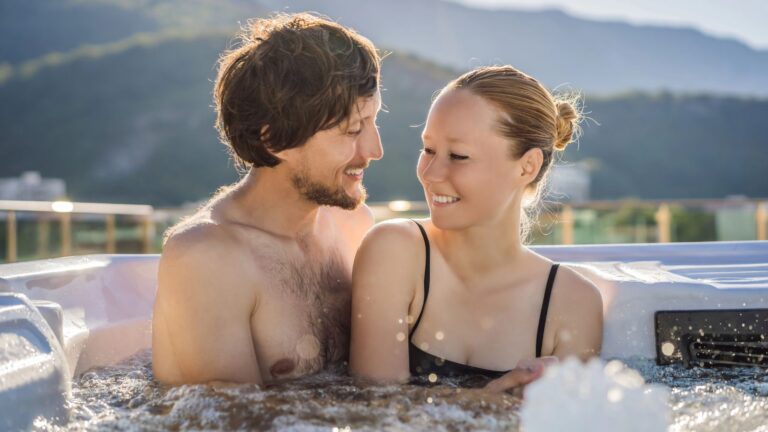 A man and woman smile at each other in an outdoor hot tub, with mountains and trees behind them.