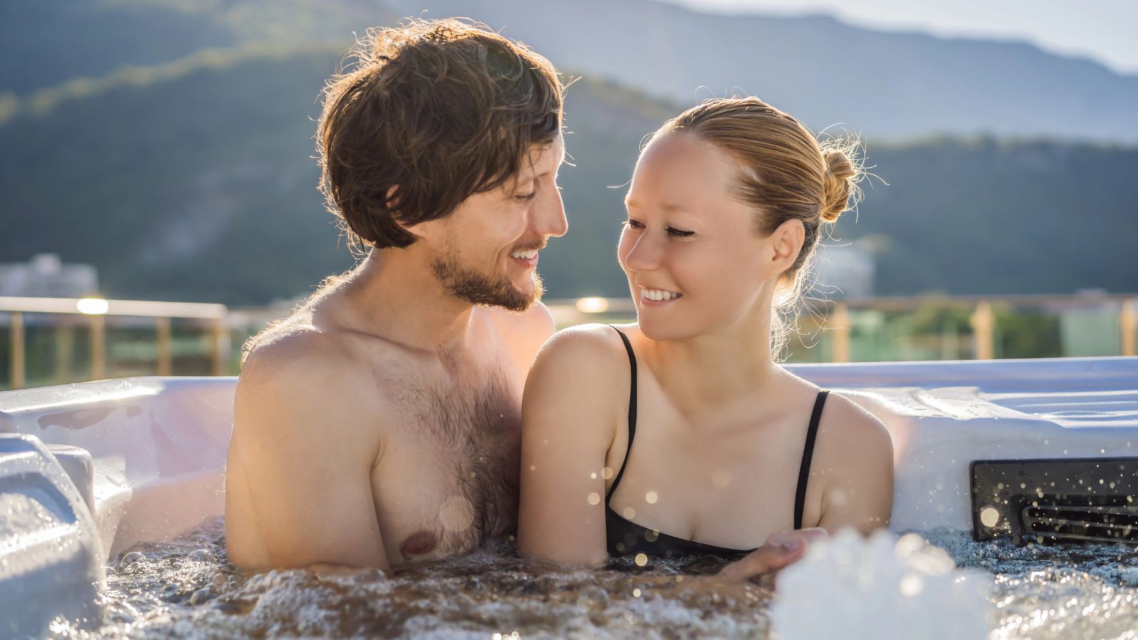 A man and woman smile at each other in an outdoor hot tub, with mountains and trees behind them.