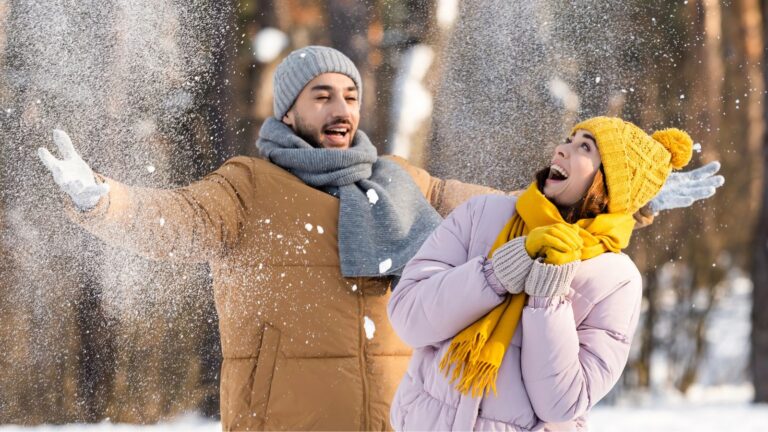 A man and woman in winter clothes stand in falling snow; he stretches his arms out while she smiles, looking upward.