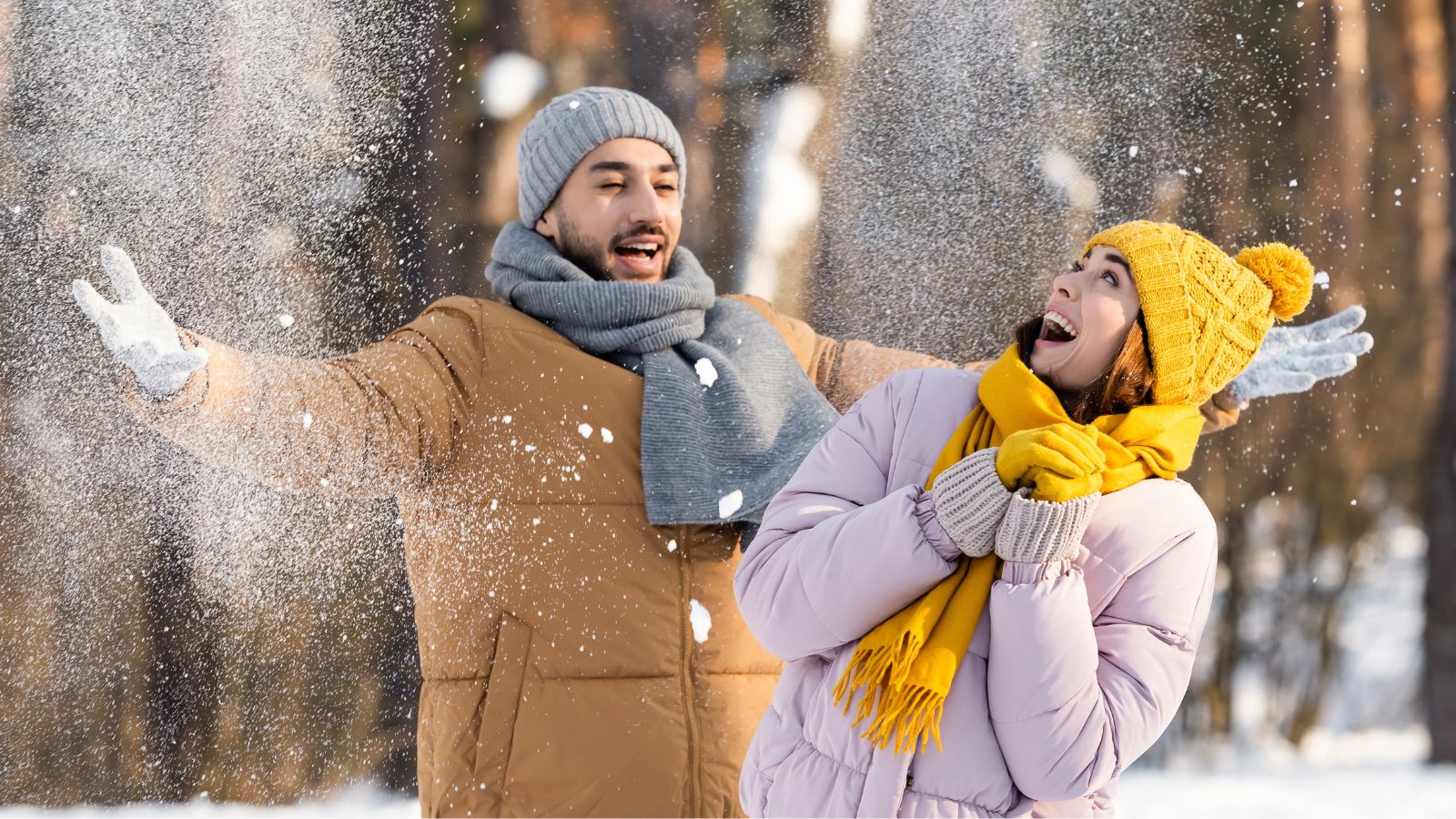 A man and woman in winter clothes stand in falling snow; he stretches his arms out while she smiles, looking upward.