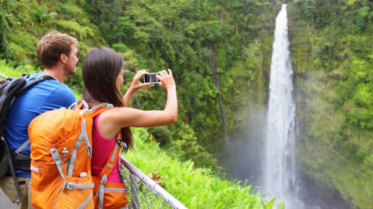 Two people with orange backpacks stand at a railing as one photographs a tall waterfall amid lush greenery.