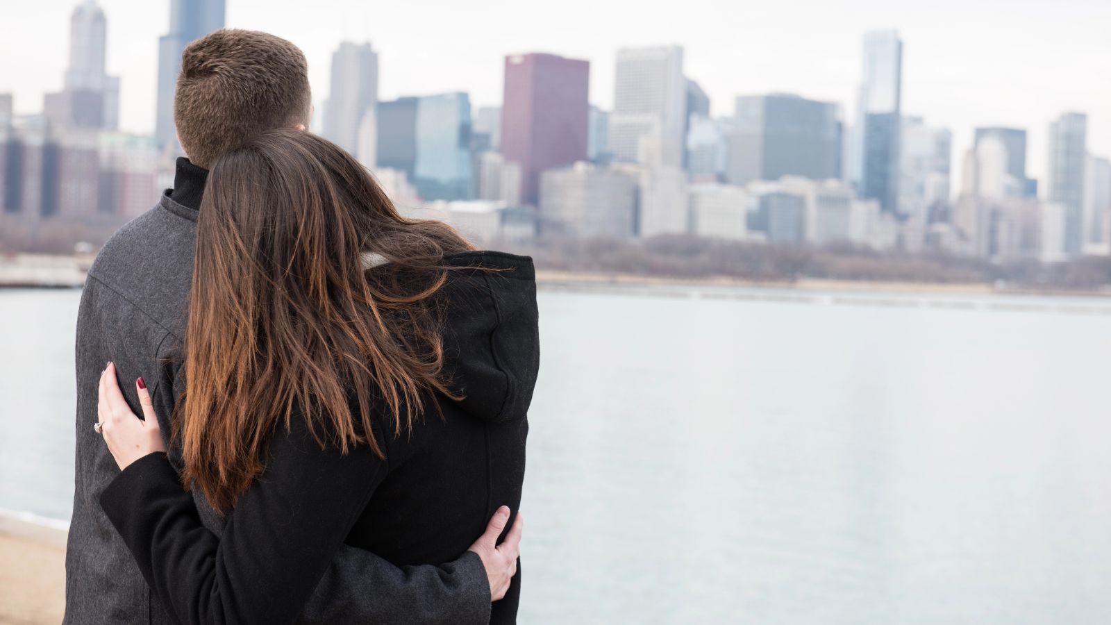 A couple embraces with their backs to the camera, gazing over water at a city skyline on a cloudy day.