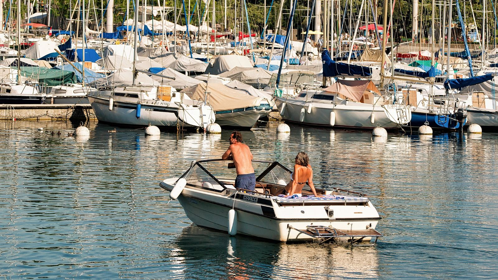 A man and woman sit on a motorboat in a sunny marina surrounded by docked sailboats and yachts.