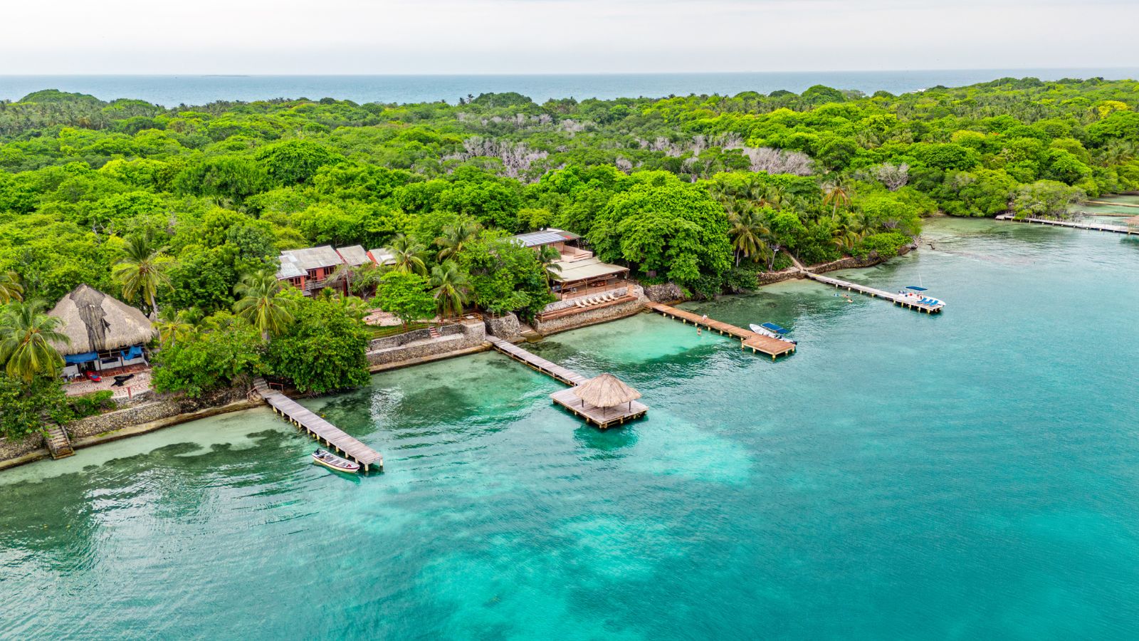 Aerial shot of a coastal resort with docks, thatched roofs, and lush greenery next to clear turquoise water.