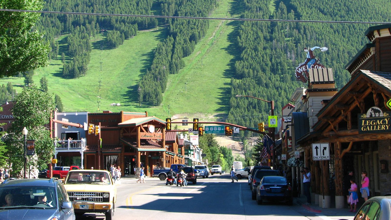 A mountain town street lined with parked cars and pedestrians, with green ski slopes rising behind the buildings.