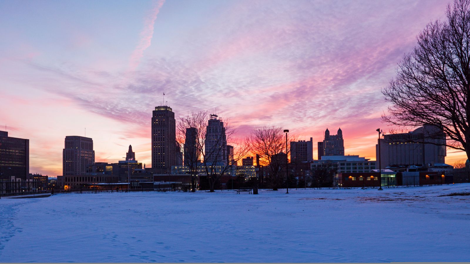 A snowy ground and bare trees frame a city skyline at sunset under colorful clouds.