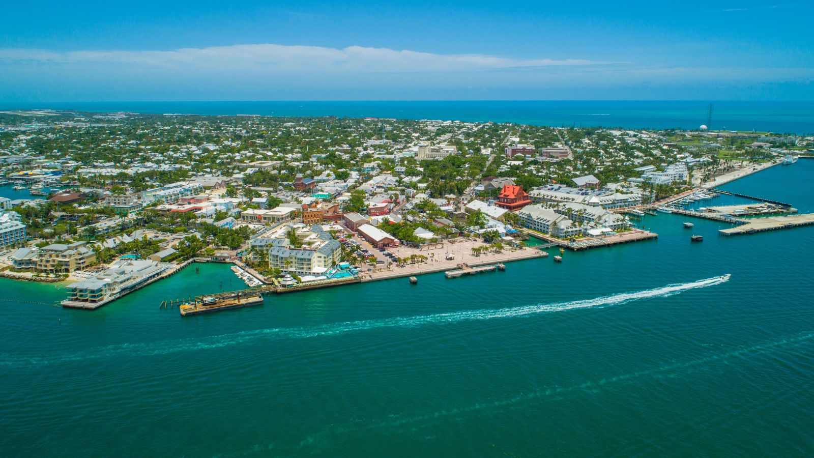 Aerial shot of a coastal city with docks, boats, and buildings by turquoise water beneath a clear blue sky.
