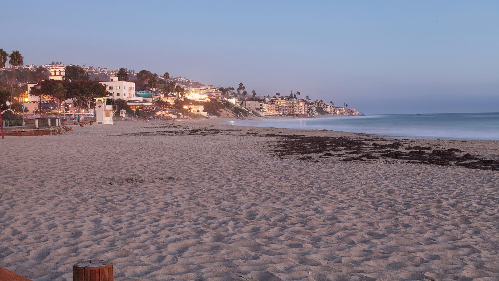 At dusk, gentle waves wash a sandy beach bordered by palm trees, coastal buildings, and glowing house lights.