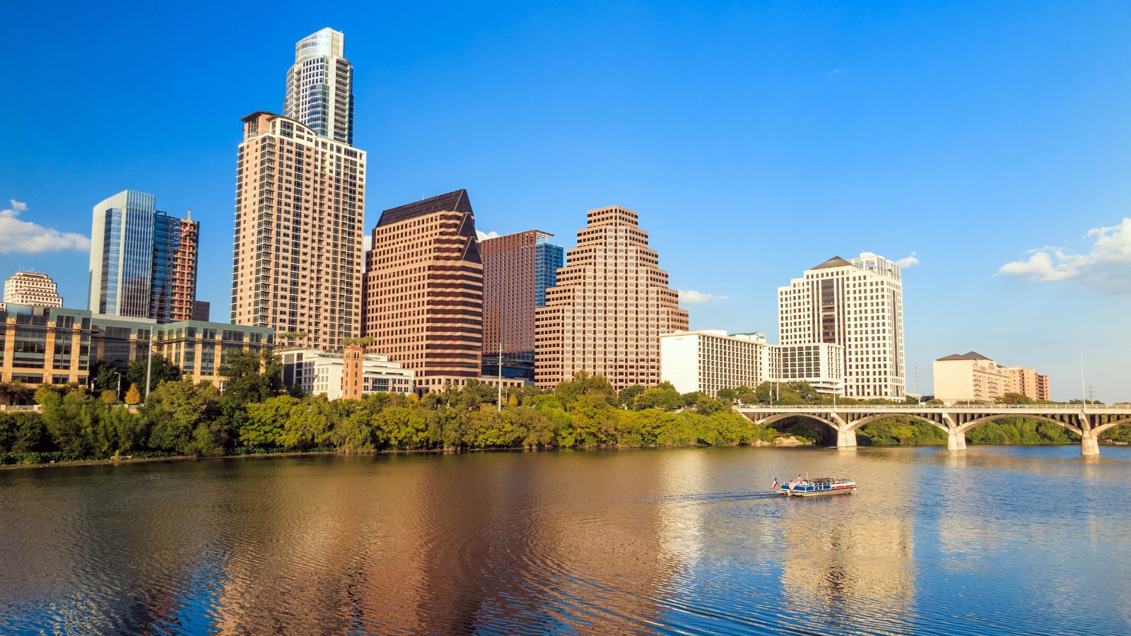 Downtown Austin skyline with modern high-rises, a river in front, and a boat on the water beneath a clear blue sky.
