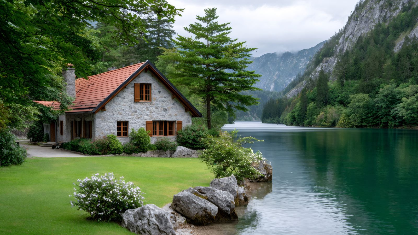 A stone house with a red roof stands beside a calm lake, with green trees, mountains, and a cloudy sky in the background.