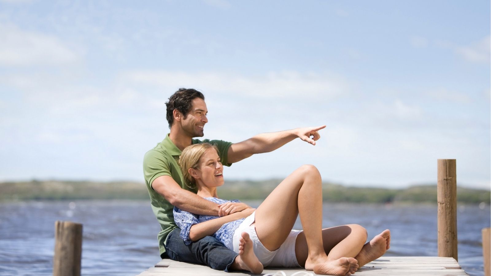 A man points while sitting with a smiling woman on a dock by water, with hills and blue sky in the background.