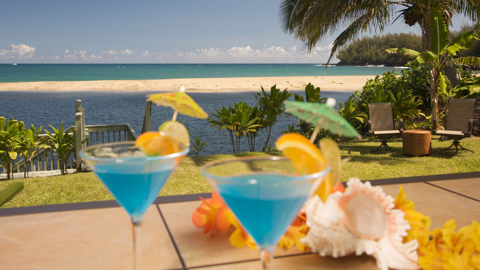 Two blue cocktails with fruit and umbrellas sit on a table facing a tropical beach with palm trees and clear water under a sunny sky.
