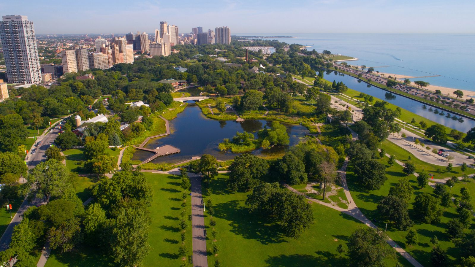 Aerial view of a city park with trees, paths, ponds, and high-rises next to a large body of water.