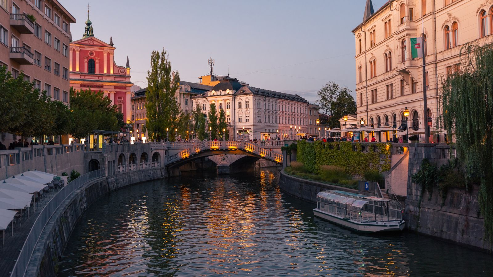 At sunset, a tour boat moves along a river through a city with historic buildings, a stone bridge, and outdoor cafes.