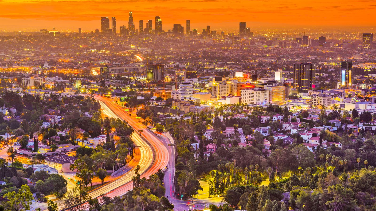 Aerial view of Los Angeles at sunset, with city lights, downtown skyscrapers, and blurred car lights on a curved highway.