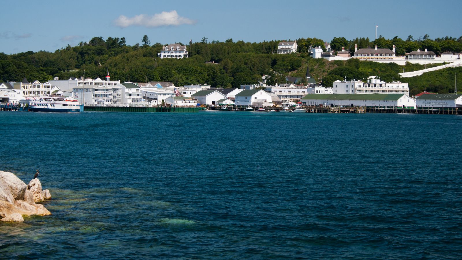 Waterfront town with white buildings, a docked ferry, and forested hills across blue water under a clear sky.