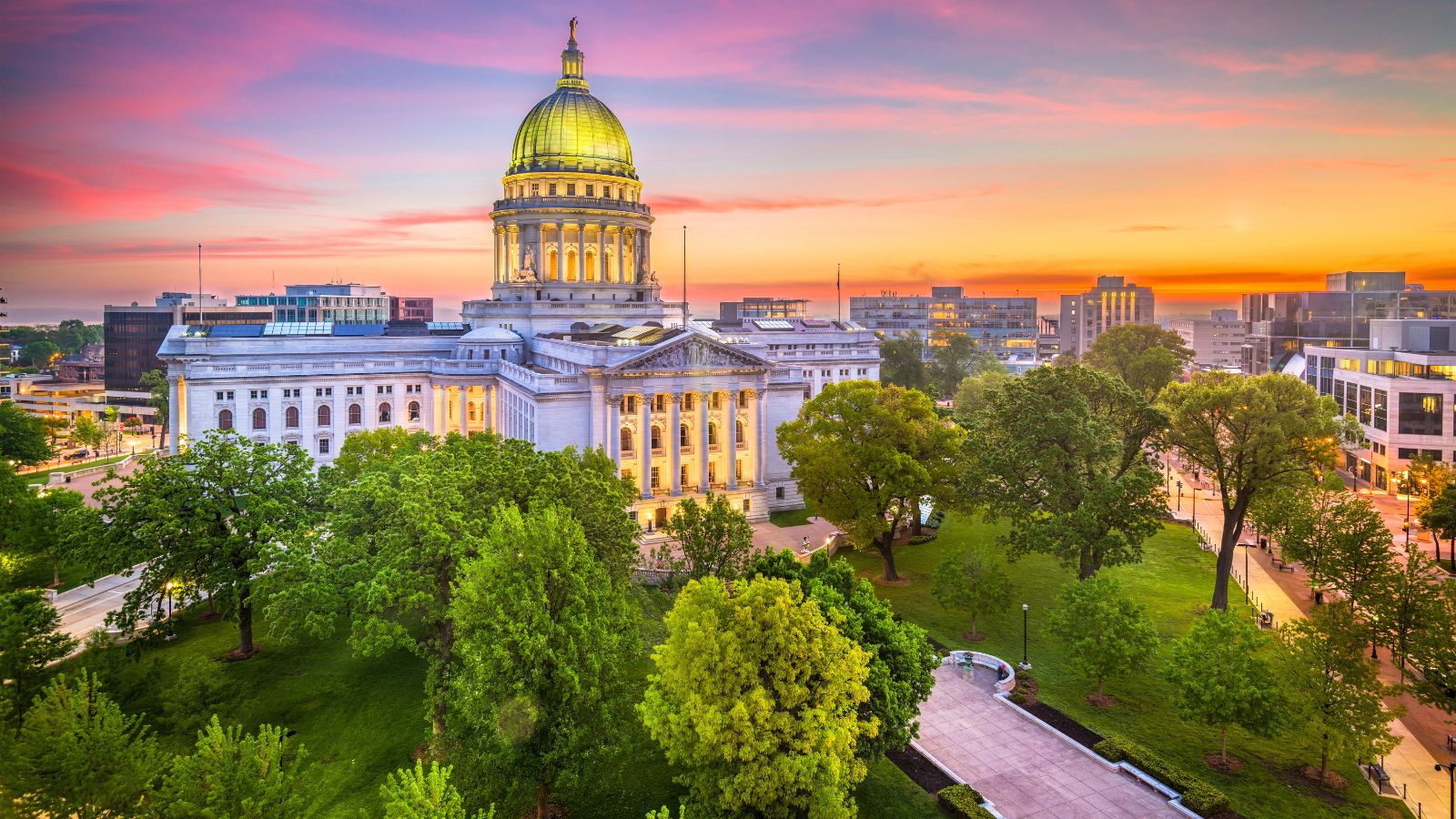 Wisconsin State Capitol with a gold dome at sunset, framed by trees and city buildings.
