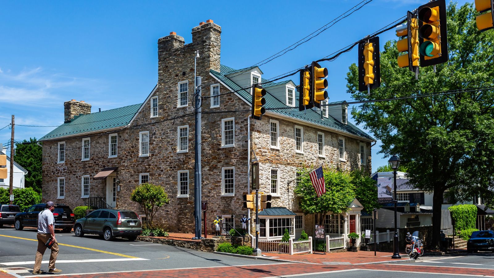 A person crosses a street by a historic stone building with two chimneys amid cars, trees, and traffic lights on a sunny day.