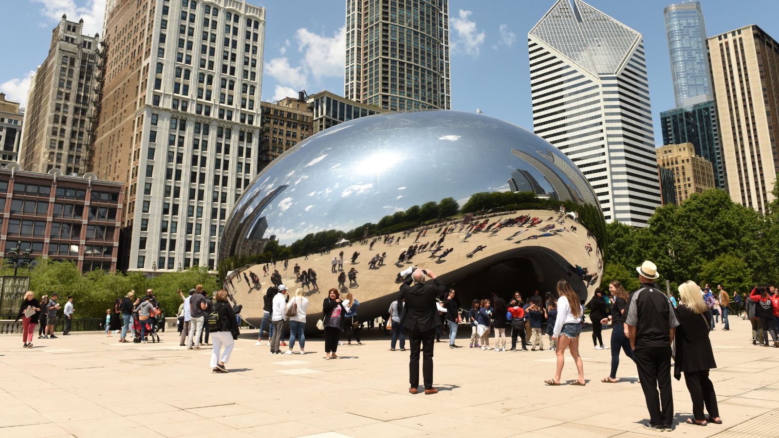 Visitors surround Cloud Gate ("The Bean") in Chicago’s Millennium Park, with skyscrapers and trees behind them.