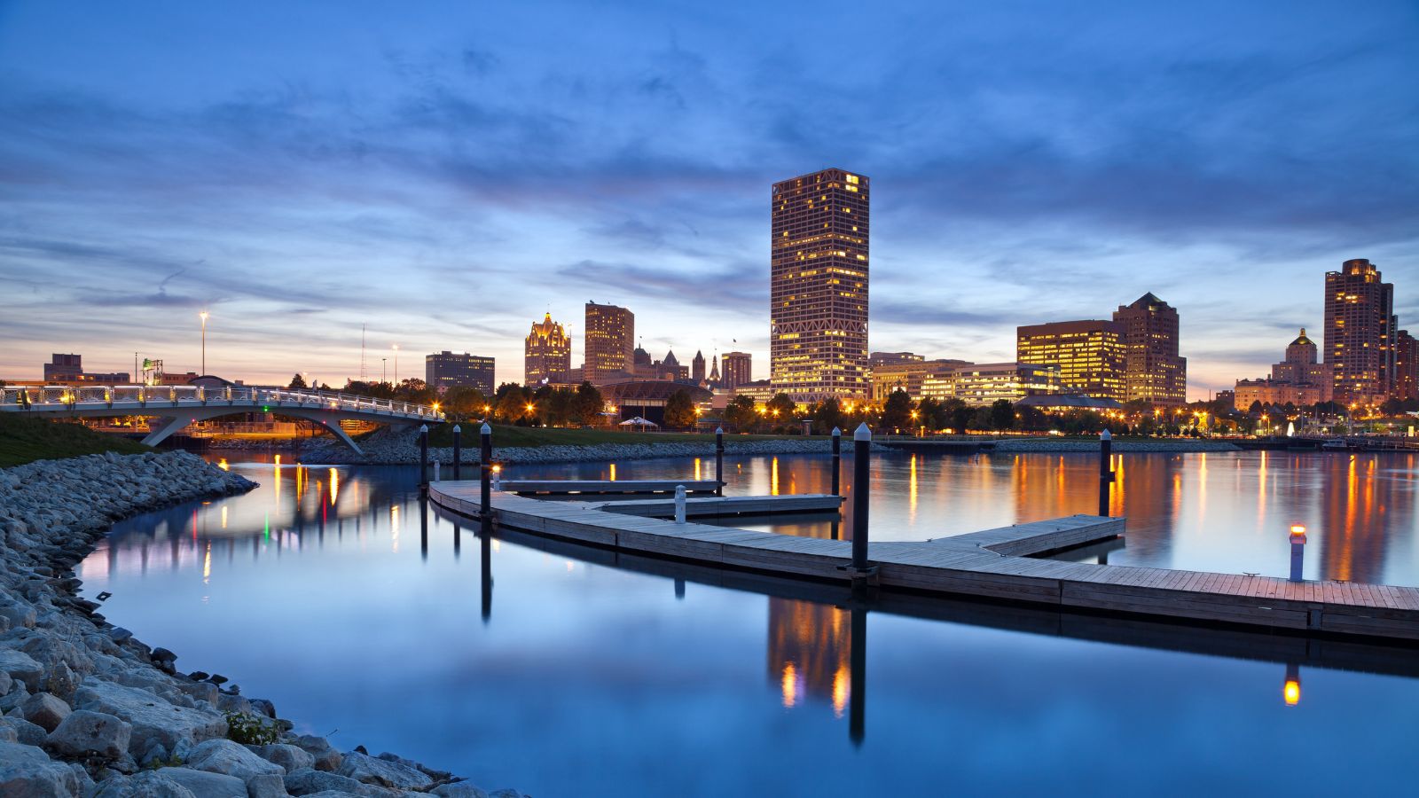 City skyline at dusk with high-rises, calm water, boat docks, and streetlights reflected on the waterfront.