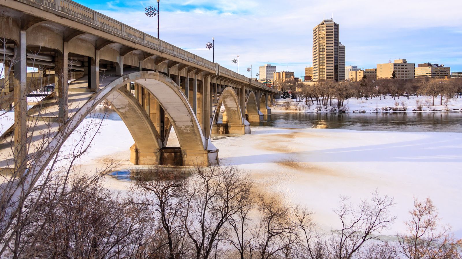 A concrete arch bridge crosses a partially frozen river, with city buildings and bare trees beneath a partly cloudy sky.