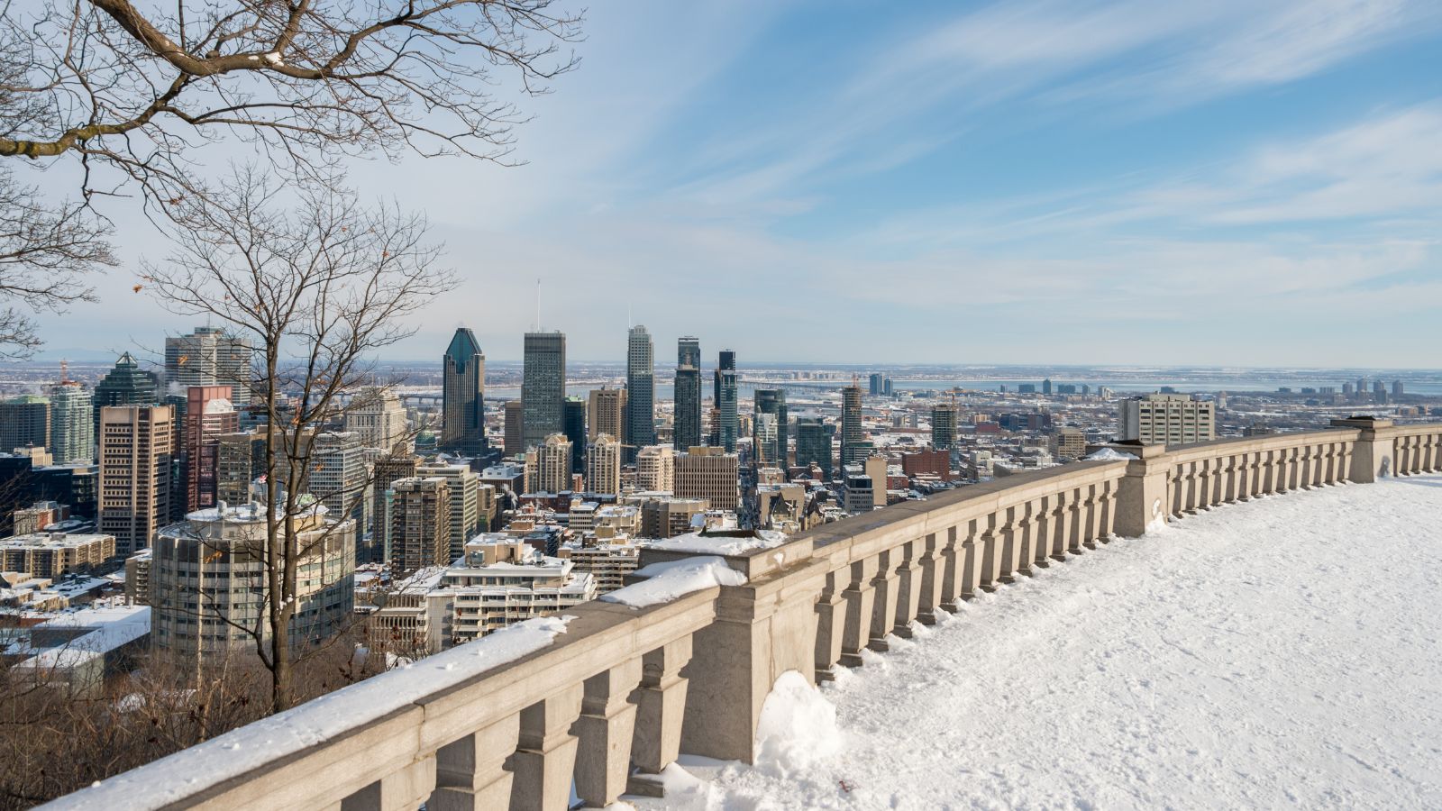 Downtown Montreal skyscrapers and buildings seen from a snowy lookout with a stone railing under partly cloudy skies.
