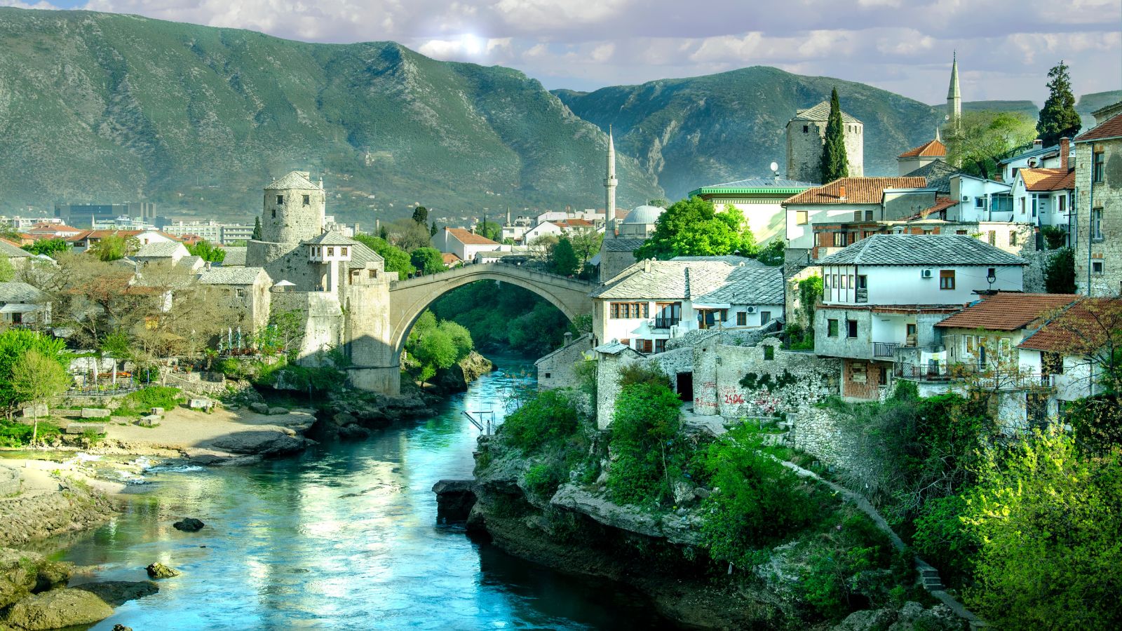 A stone arch bridge crosses a river in a historic town with stone buildings and mountains in the background.