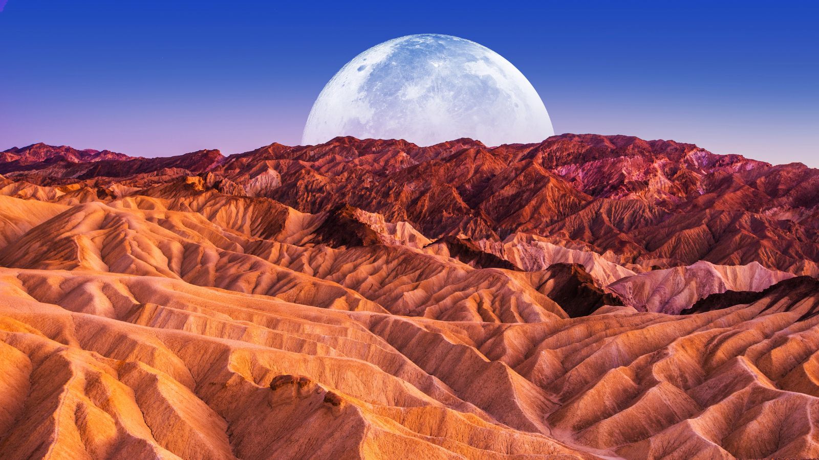 Colorful desert hills with sharp ridges, distant rugged mountains, and a full moon rising in a clear blue sky.