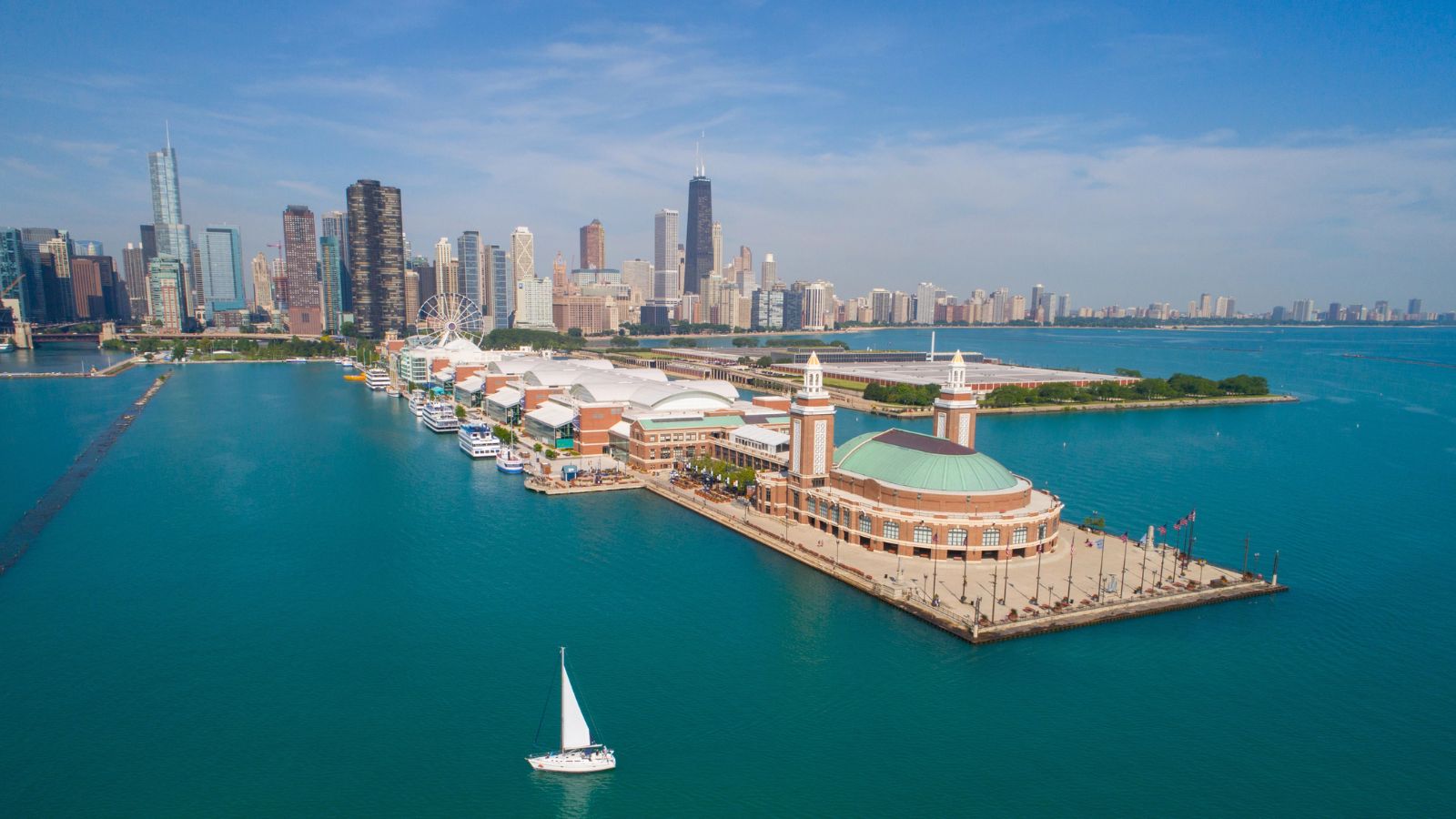 Aerial view of Navy Pier jutting into Lake Michigan, with Chicago’s skyline behind and a sailboat on the water.