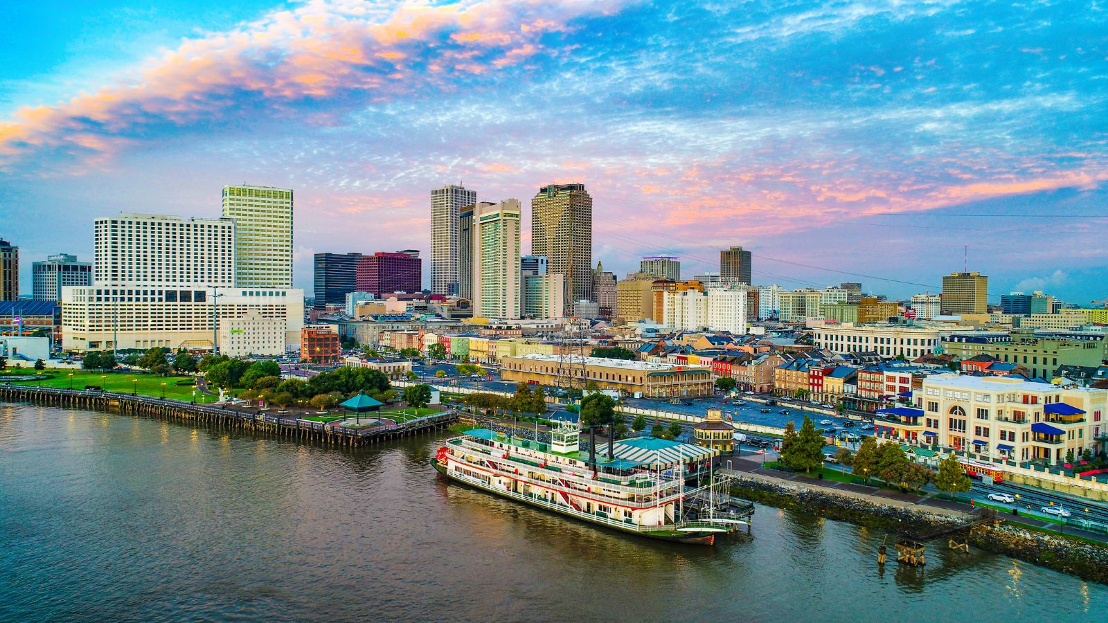 Aerial view of downtown New Orleans with skyscrapers, historic buildings, and a riverboat on the Mississippi River at sunset.