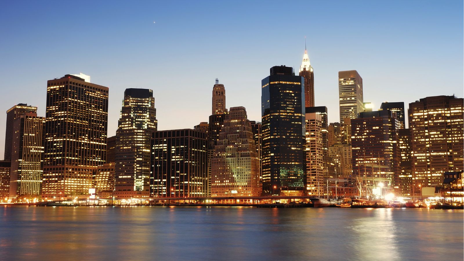 Dusk city skyline with lit skyscrapers reflecting on calm water beneath a clear evening sky.
