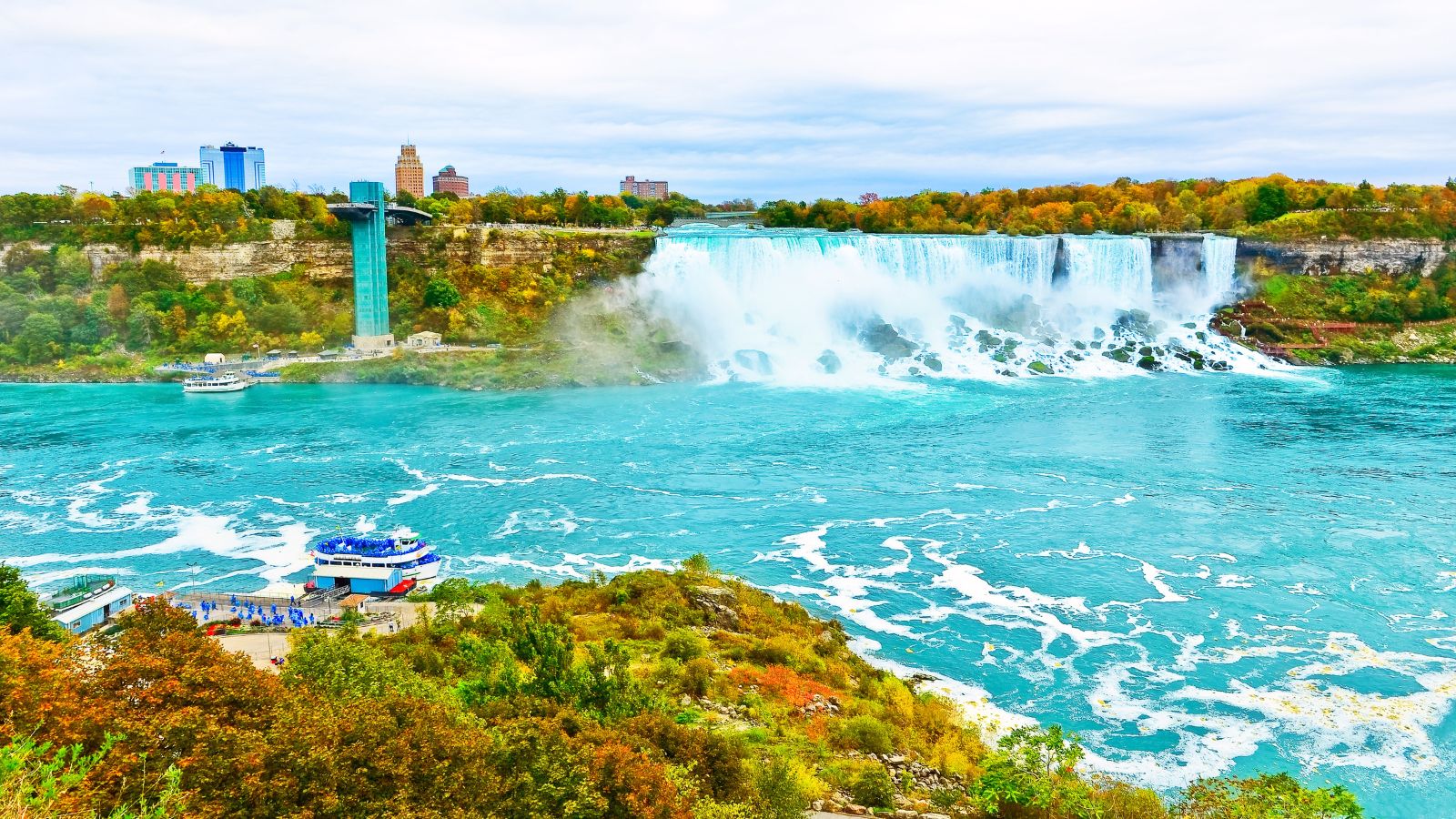 Niagara Falls with waterfalls, observation tower, tour boat on turquoise river, and city buildings under an overcast sky.